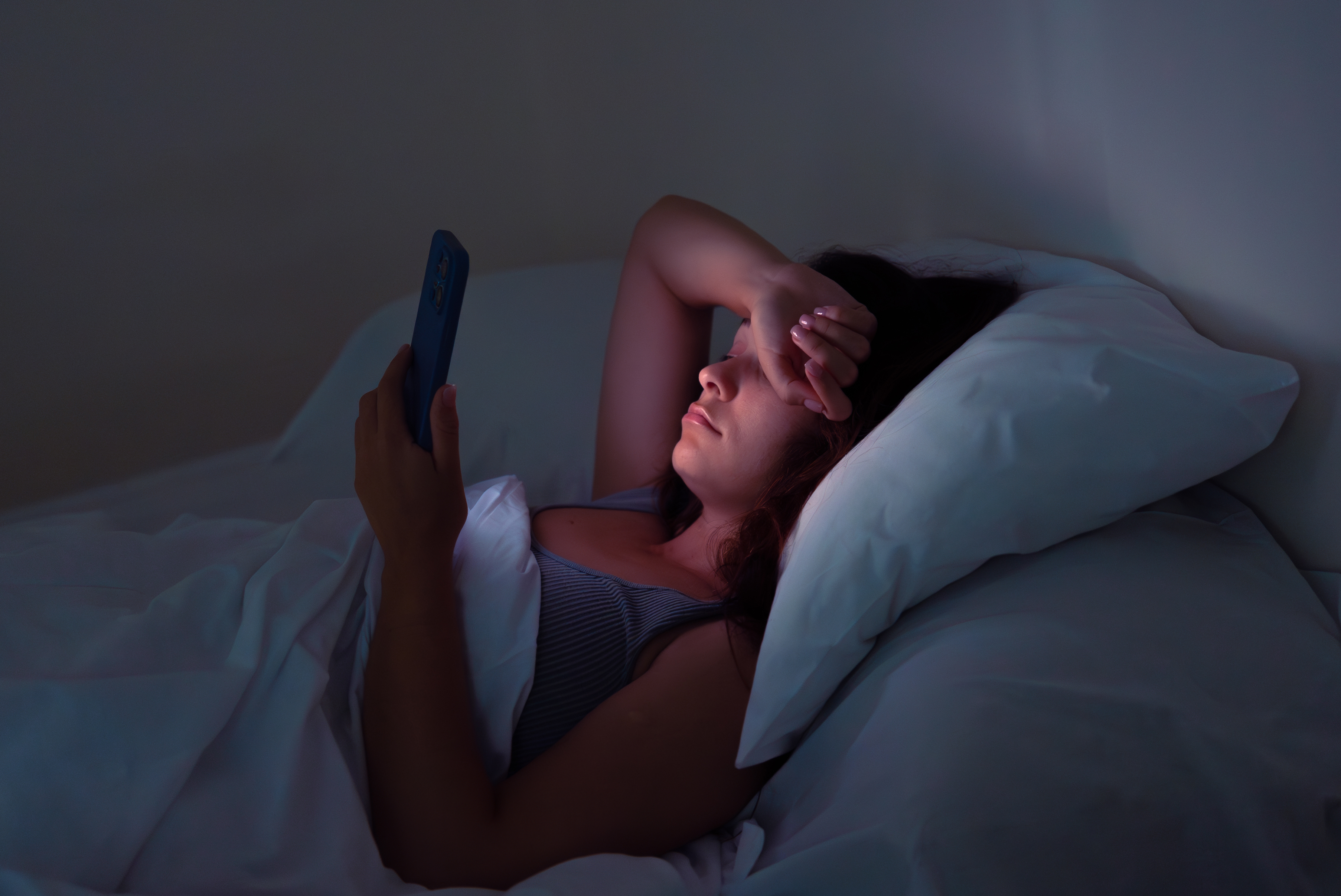 Tired young woman browses information on smartphone before sleep lying in bed in semi-dark bedroom